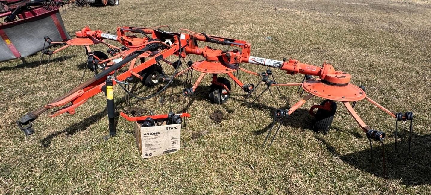 An orange rotary hay rake sits in a grassy field, its arms folded in an upright transport position.