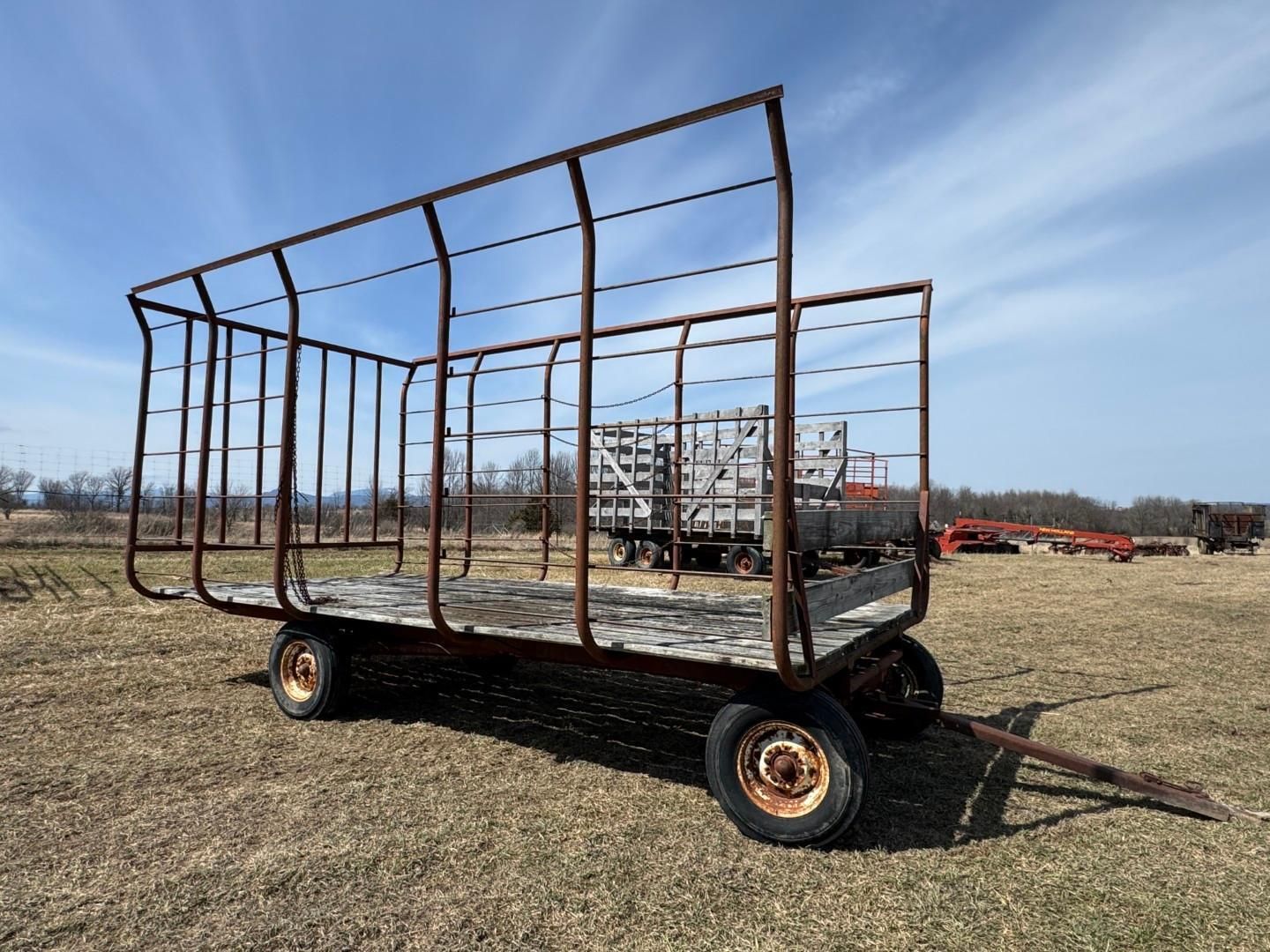 An empty metal-framed farm wagon with a flat wooden bed sits in a grassy field under a blue sky.