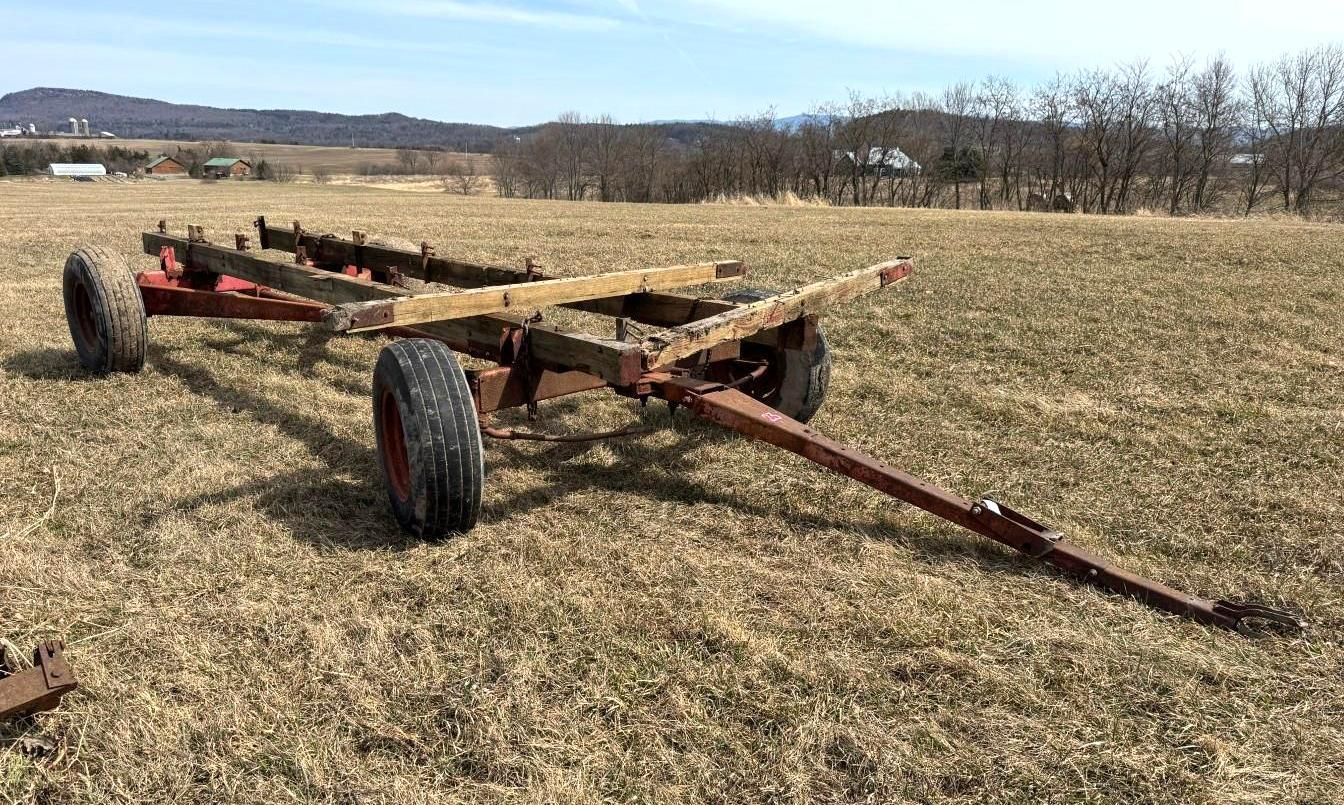 A red-painted farm wagon chassis with wooden beams sits in a grassy field under a bright sky.