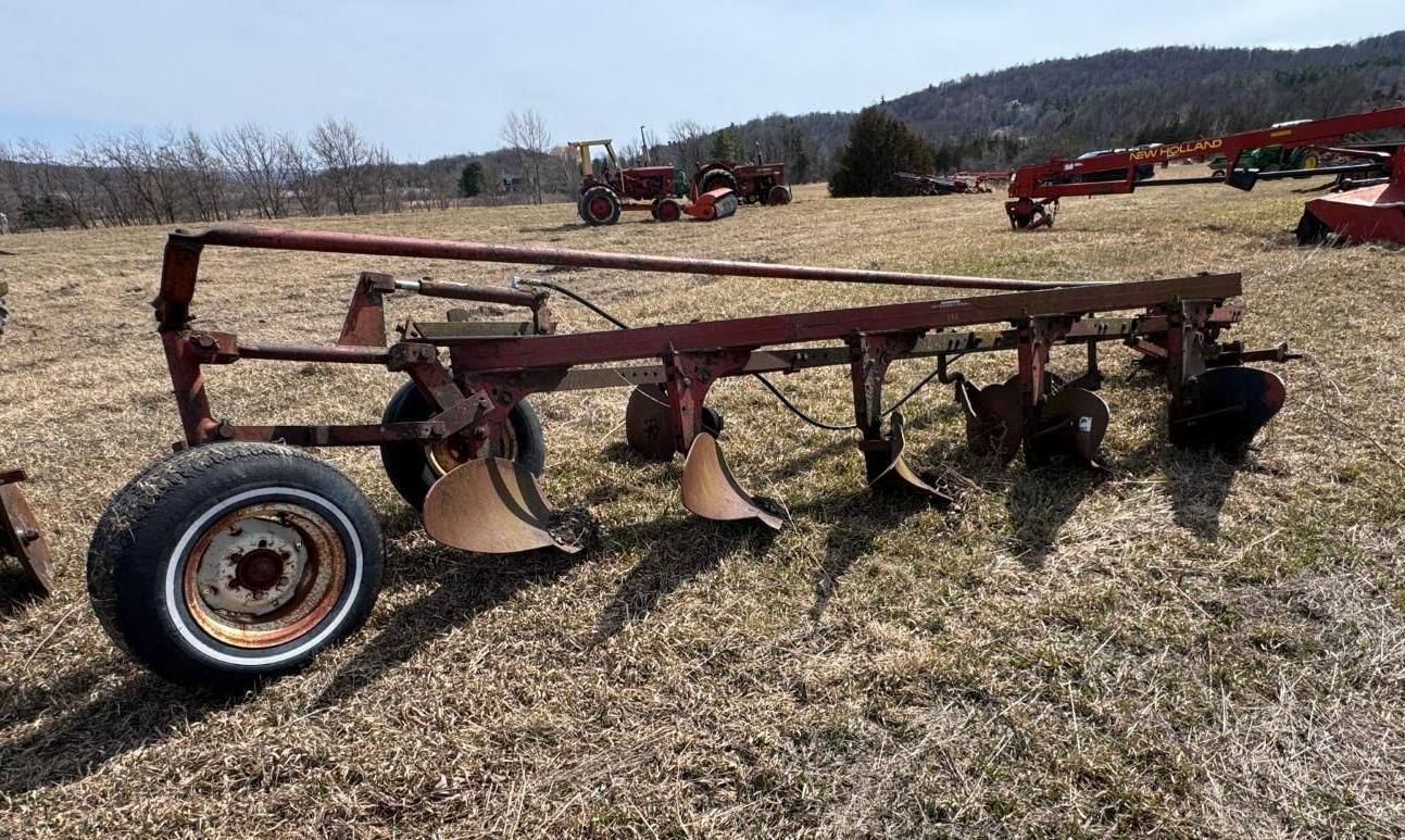 A red, pull-type moldboard plow with multiple bottoms and a single support wheel, sitting in a grassy field.