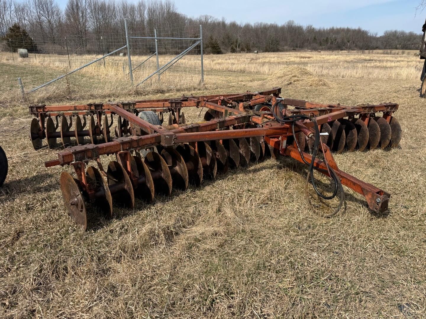 An orange heavy-duty agricultural disc harrow sits in a grassy field under a clear blue sky.