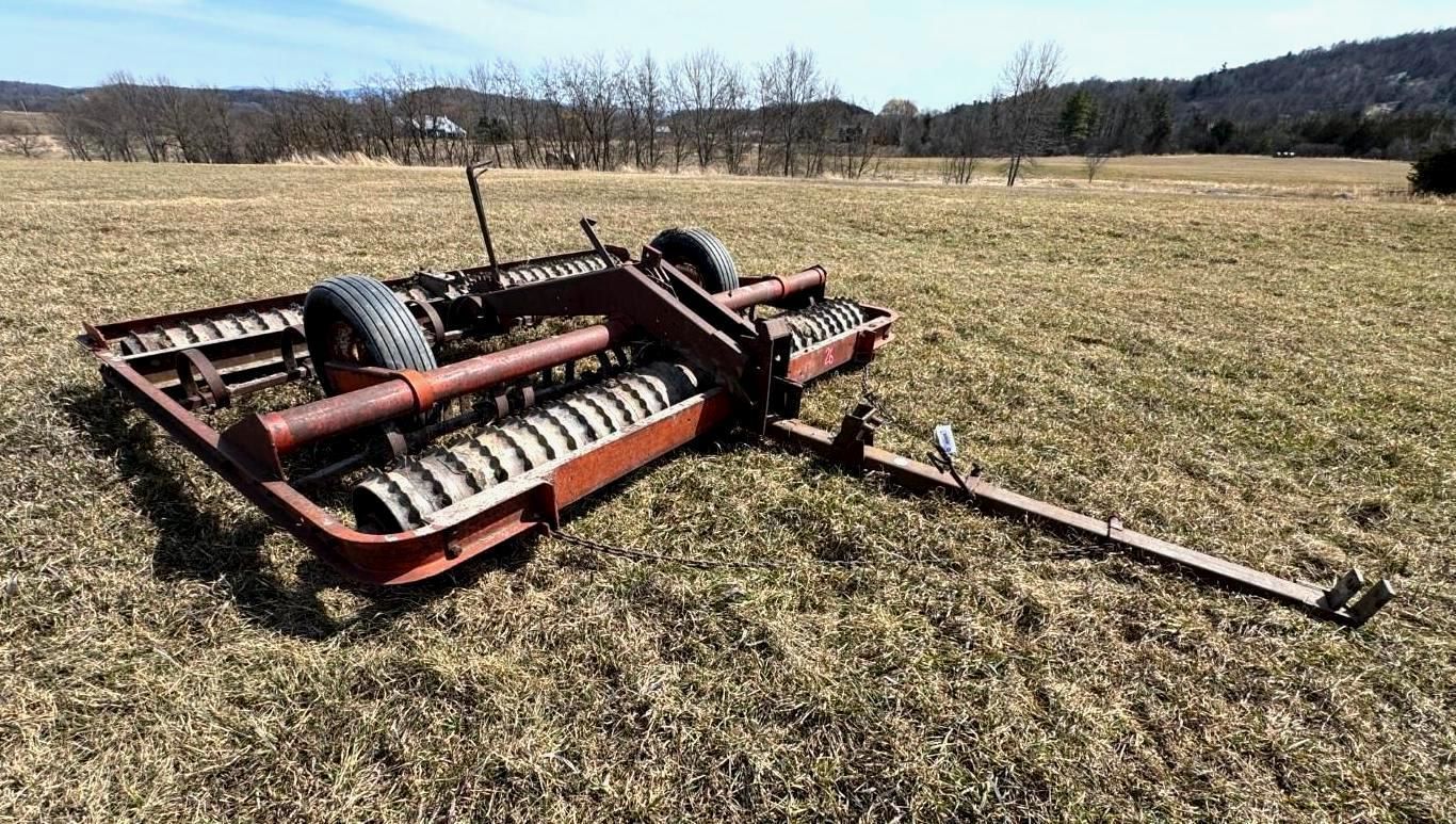 A red cultipacker agricultural implement sitting in a grassy field under a clear blue sky.