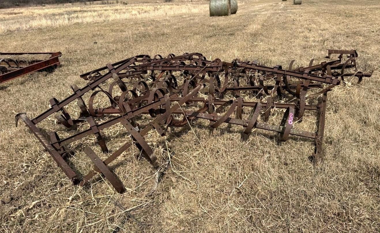 A multi-row farm cultivator resting in a harvested field with hay bales in the background.