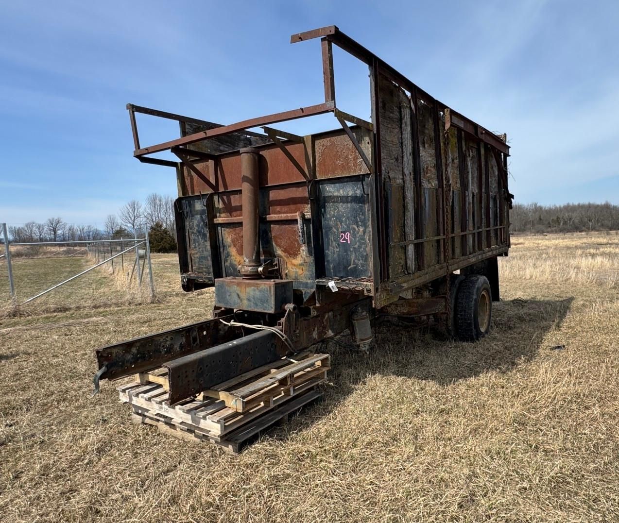 A metal dump trailer or farm wagon stands in a grassy field under a clear blue sky.