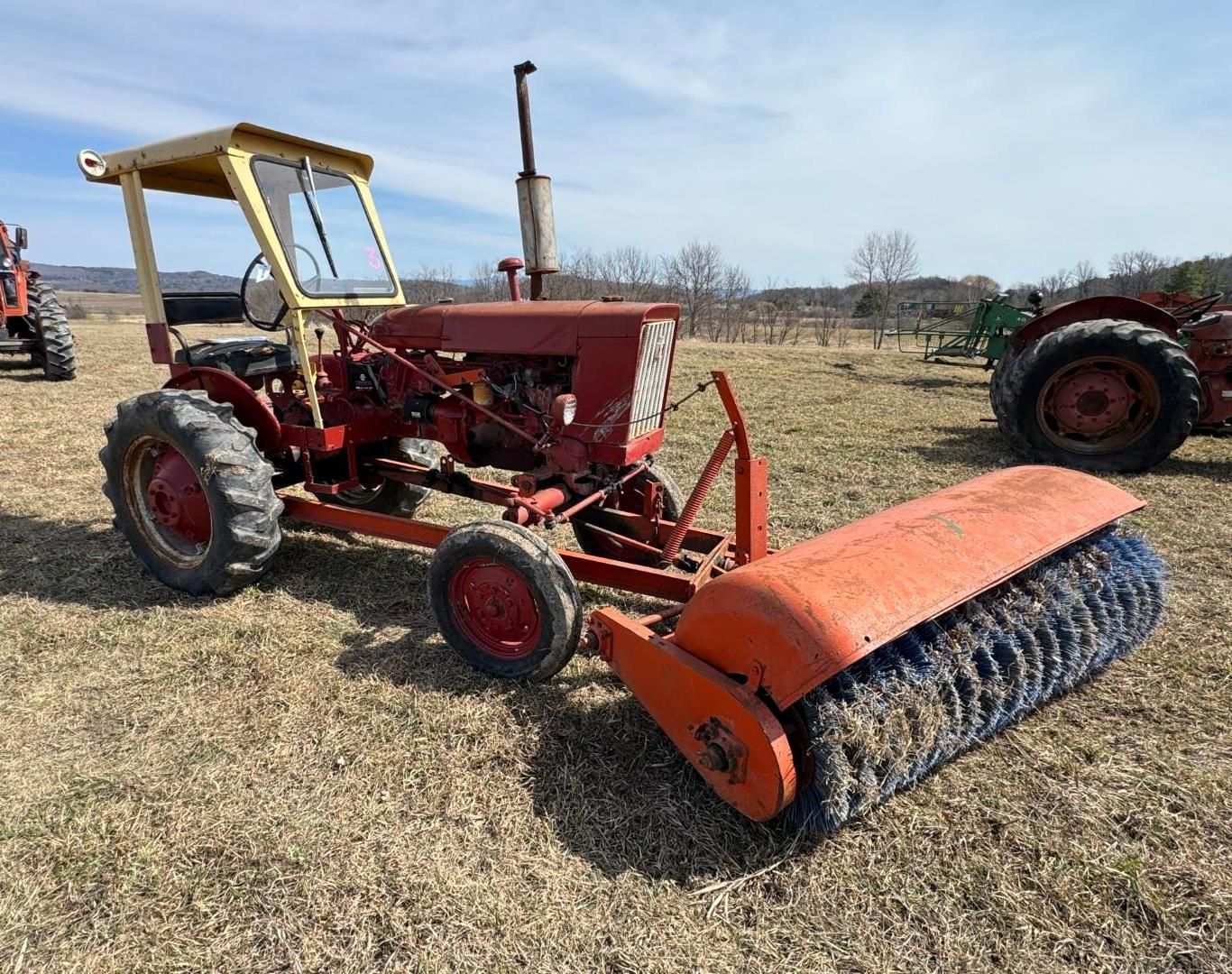 A red tractor with a large, front-mounted industrial broom attachment sits in an open field under a blue sky.