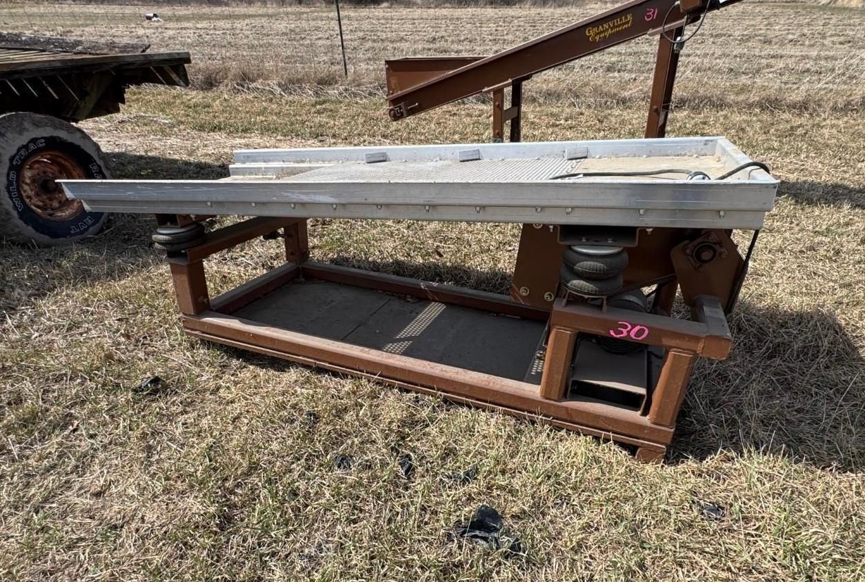 A vibrating sorting table with a metal top and brown steel frame, positioned outdoors on a grassy field.