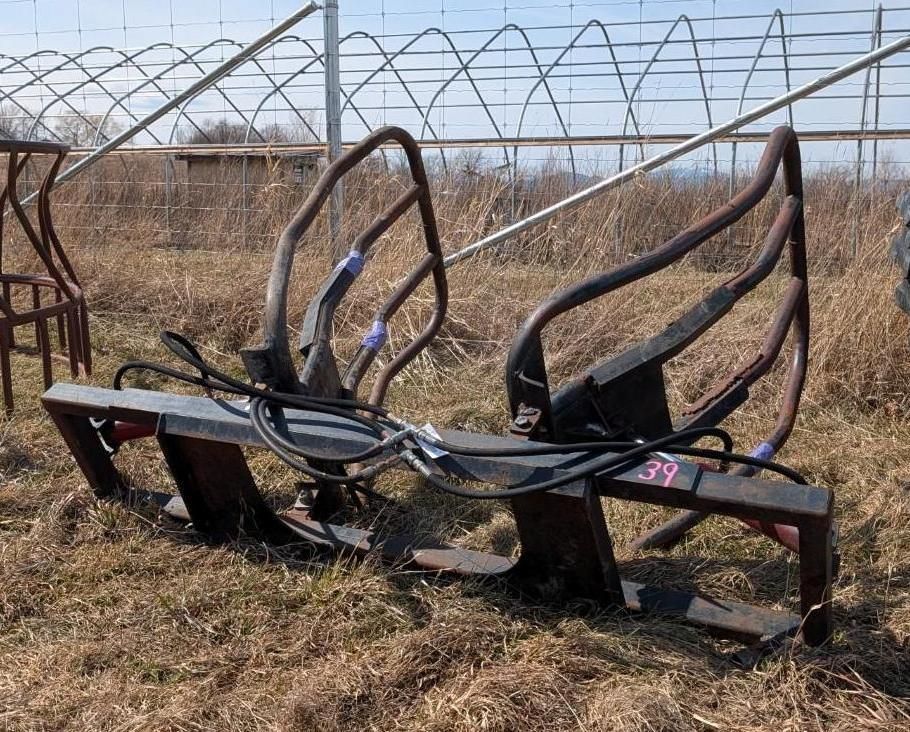 A black, hydraulic bale grab attachment with two curved metal gripping arms sitting in a grassy field.