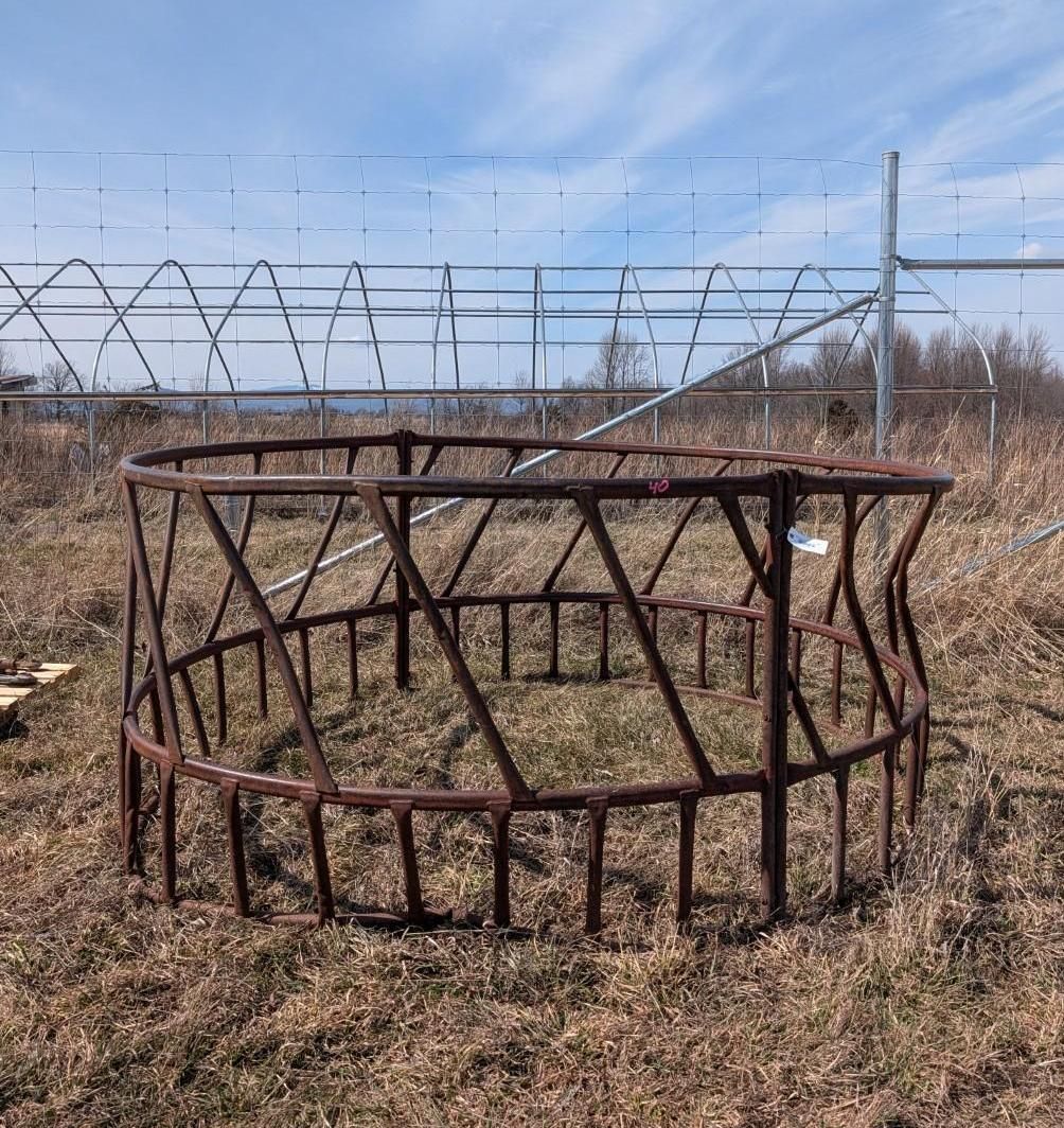 A circular metal hay feeder sits in a field in front of a metal-framed agricultural structure.