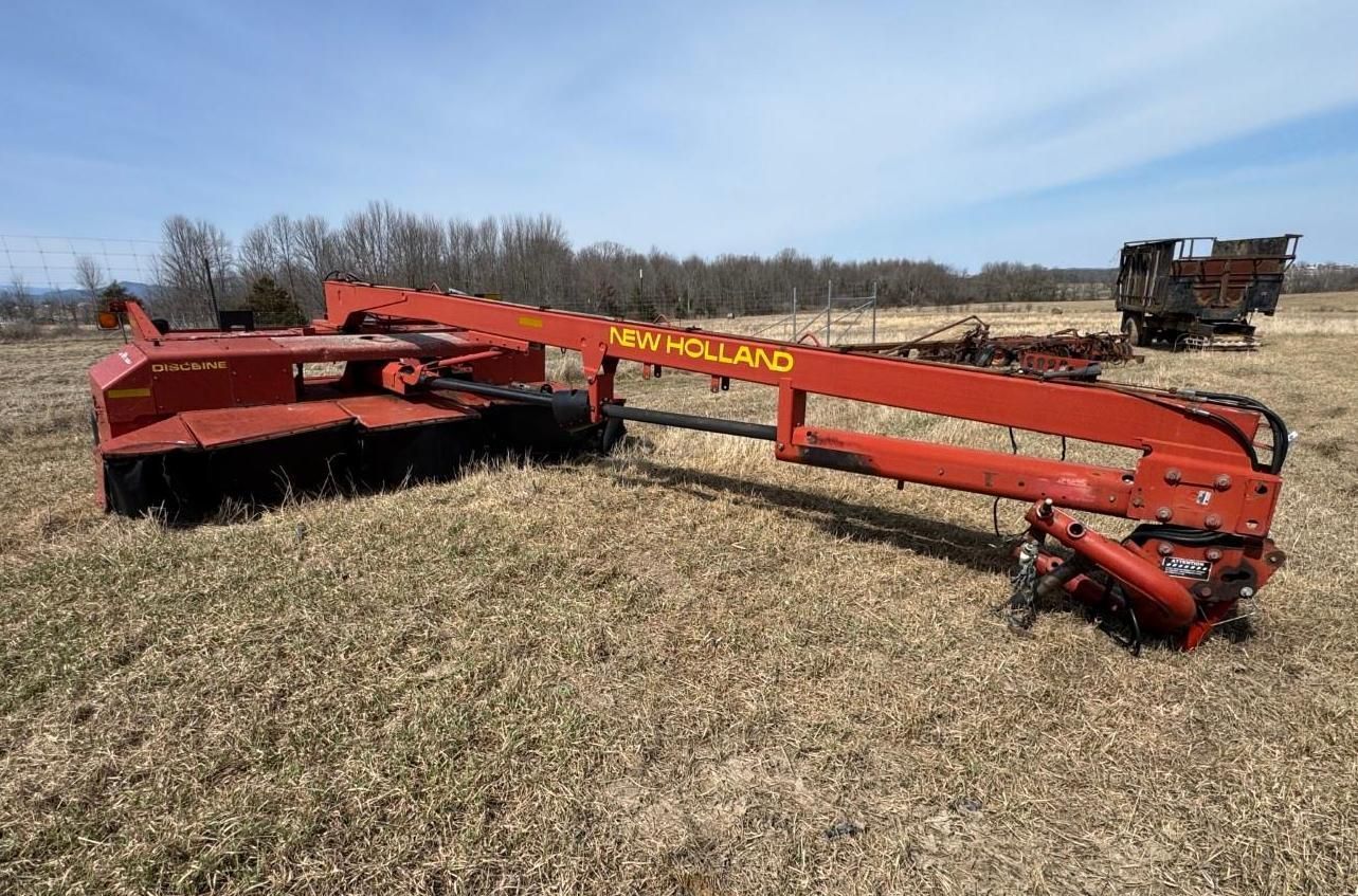 An orange New Holland disc mower conditioner sitting in a field on a sunny day.