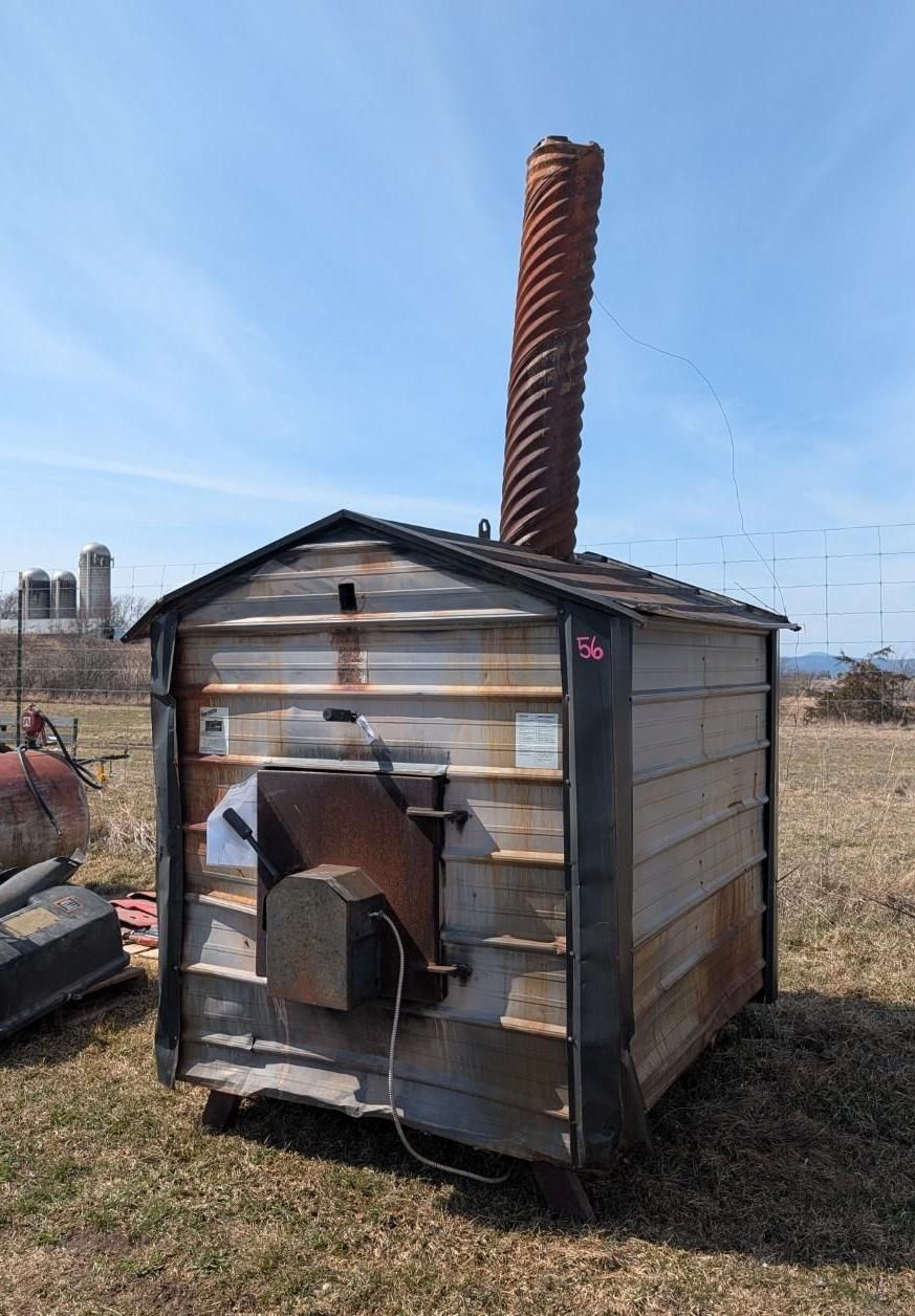 An outdoor wood sugar shack with a spiral-textured chimney, enclosed in a rustic wooden shed in a grassy field.