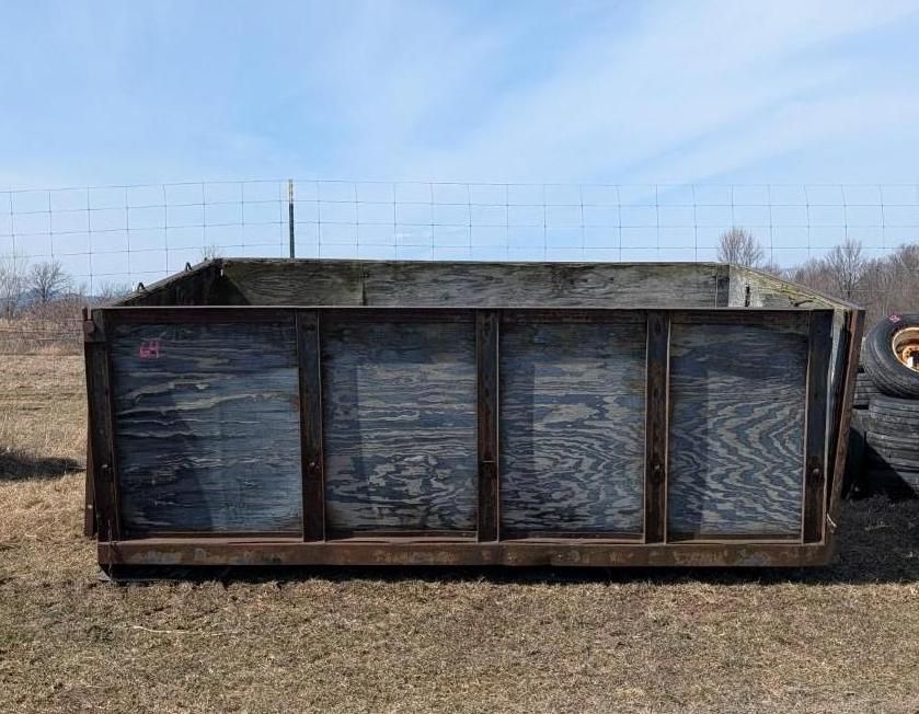 A rectangular, open-topped metal trailer frame with dark wood panel sides sits on a grassy field under a sunny sky.