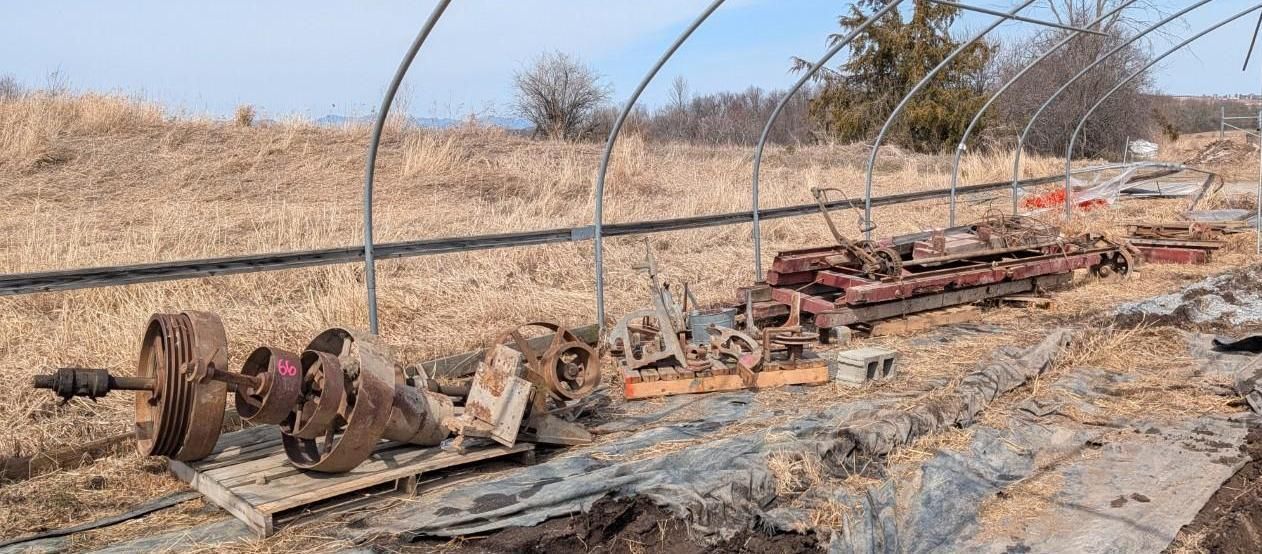 Farm equipment and steel metal parts lay on the ground inside a long, open hoop house frame in a field.