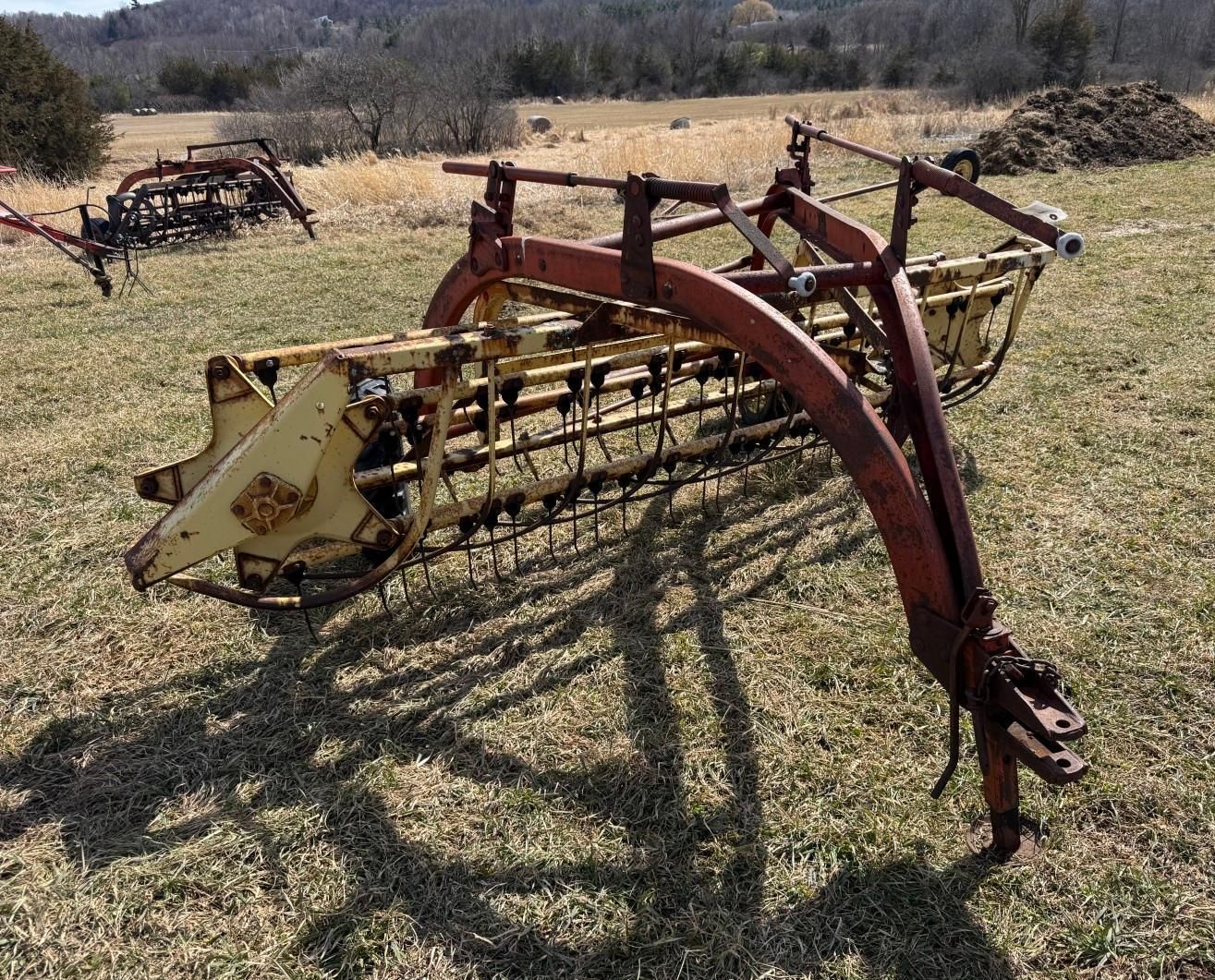 A vintage horse-drawn hay rake with a red metal frame and yellow rotating tines, sitting in a grassy field.