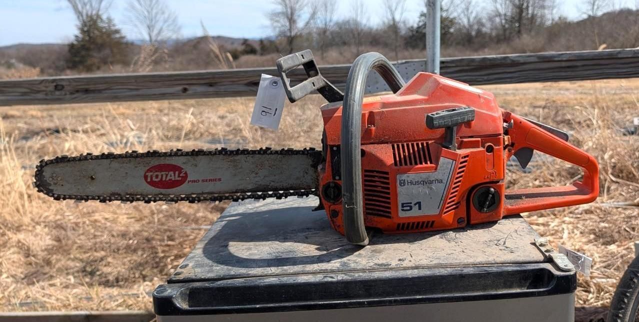 A bright orange chainsaw sits on a gray surface in an outdoor, grassy field.