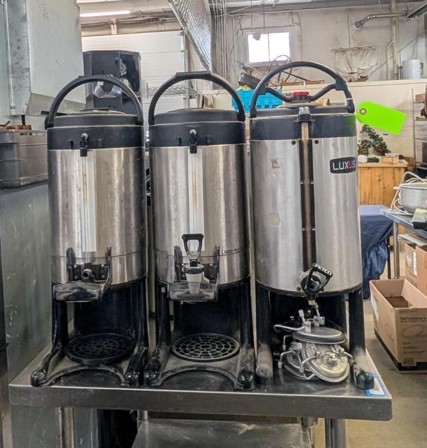 Three stainless steel beverage dispensers on a cart in a workshop or storage area