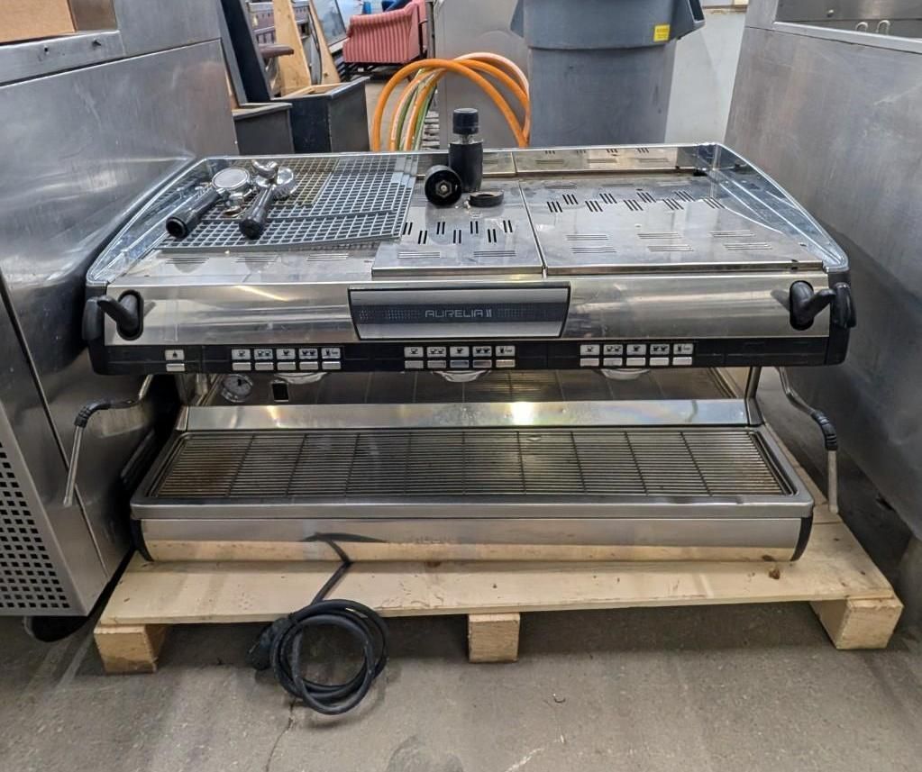 Industrial food processing machine on a wooden pallet in a workshop, with metal trays and black housing