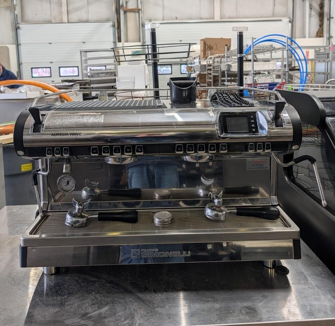 Stainless steel espresso machine on a counter in a workshop, with gauges, portafilters, and blue hoses.