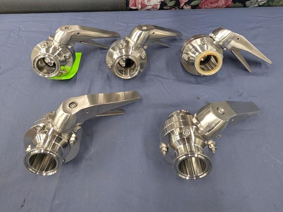 Five shiny metal pipe fittings displayed on a blue cloth, with one on a green tag and one with a brass-colored ring