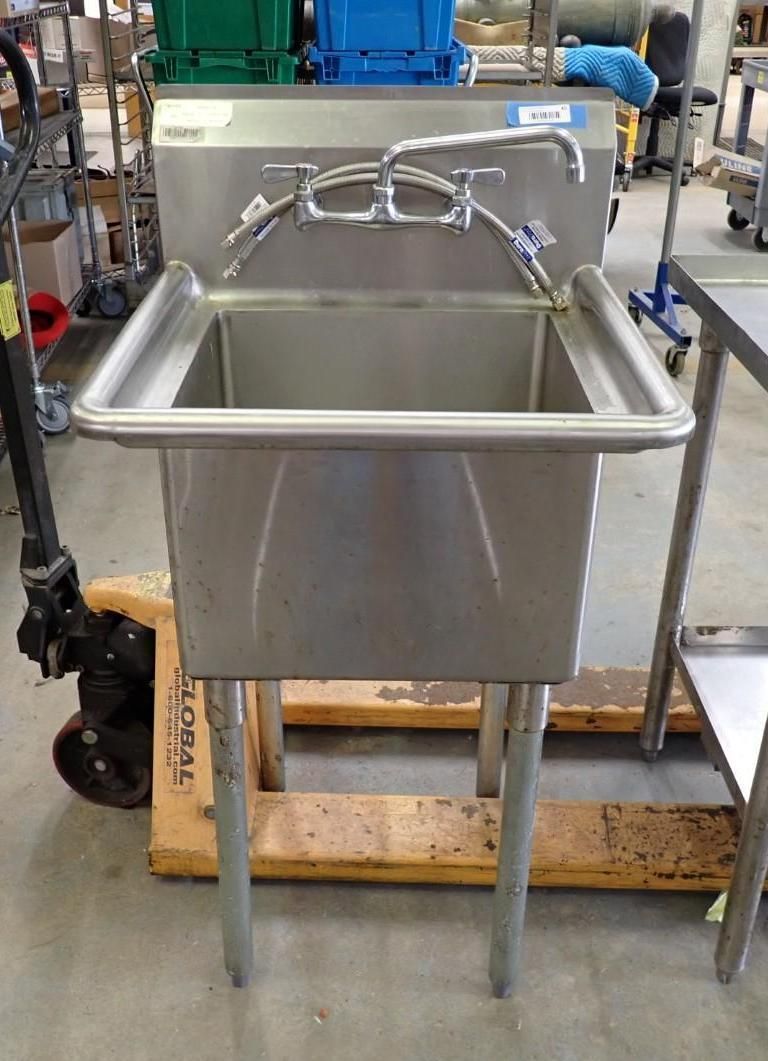 Stainless steel industrial sink with dual faucets on a wooden pallet in a workshop or warehouse.
