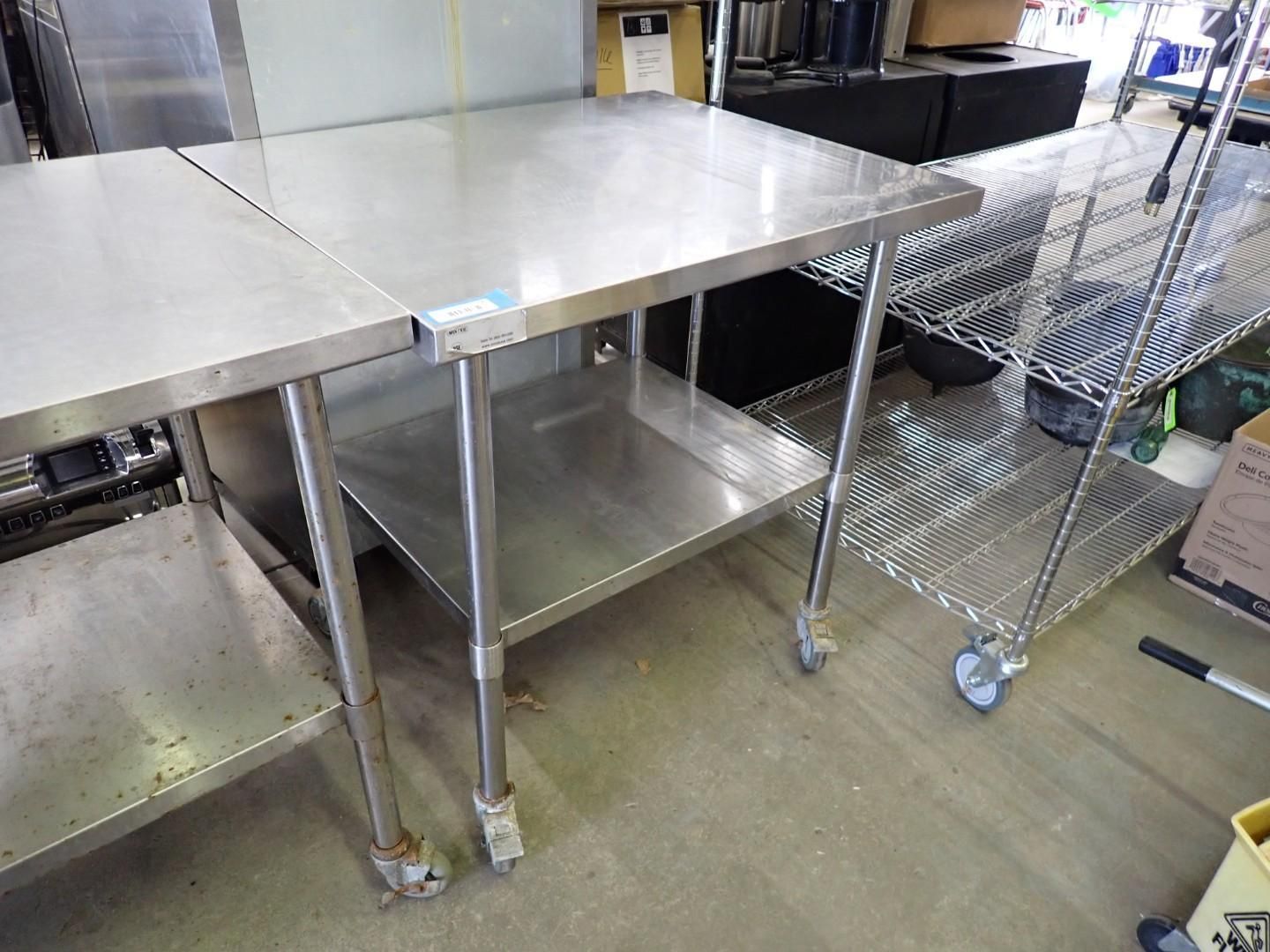 Stainless steel worktables in a commercial kitchen or prep area, with mesh carts and shelves nearby.