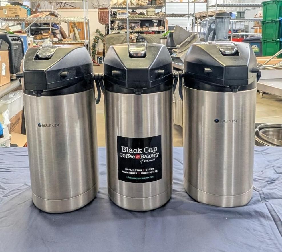 Three stainless steel coffee urns on a table in a warehouse setting, with the center urn labeled Black Cap Coffee Co.