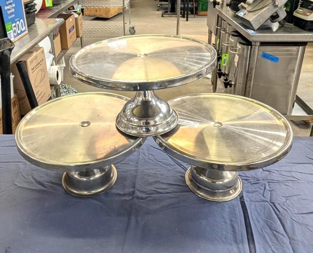 Three-tier chrome cake stand with round glass shelves on a blue cloth in a shop.