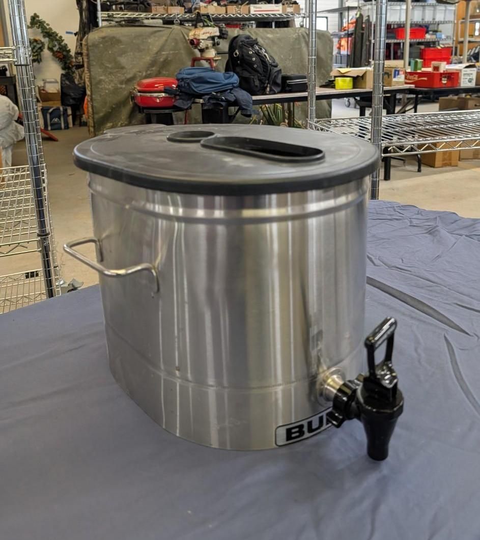 Stainless steel beverage urn with black lid and spigot on a table in a warehouse-like room.