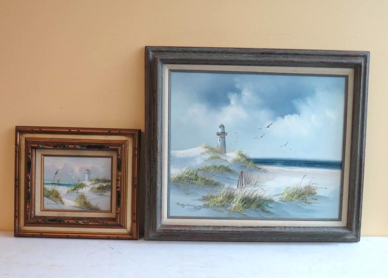 Two framed paintings: one large, one small. Both depict a sandy beach scene with a lighthouse under a cloudy sky.