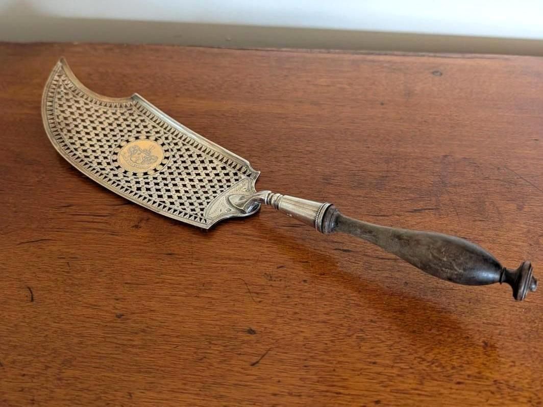 A silver fish slice with a pierced, ornate blade and a dark, shaped handle, resting on a polished wooden surface.