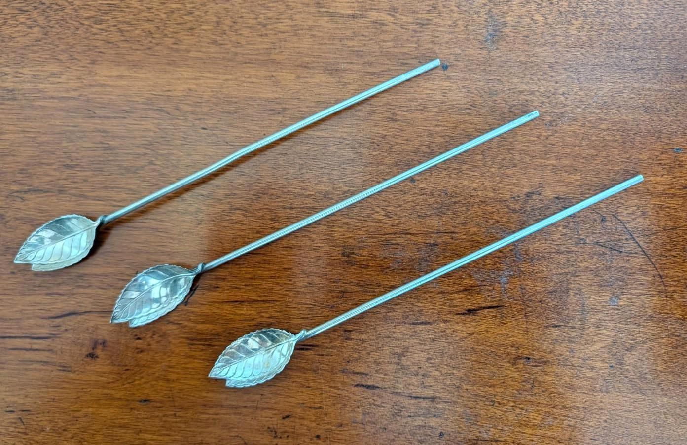 Three silver-colored cocktail picks with leaf-shaped ends, arranged diagonally on a wooden surface.