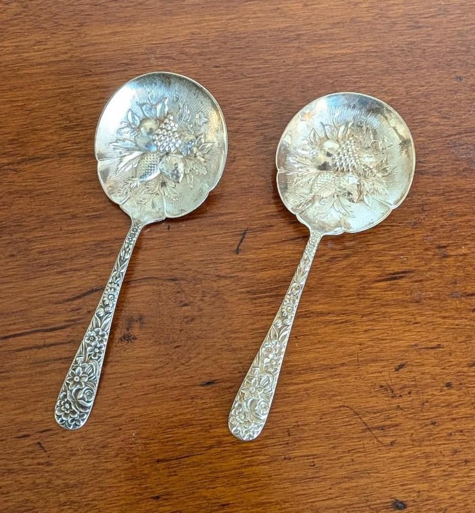 Two silver spoons with intricate floral engravings in the bowls and on the handles, resting on a dark wooden surface.