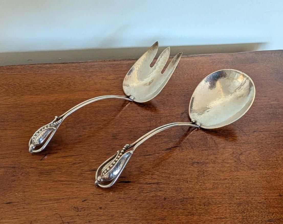 A pair of polished silver-toned salad serving utensils with ornate, textured handles, resting on a dark wood surface.