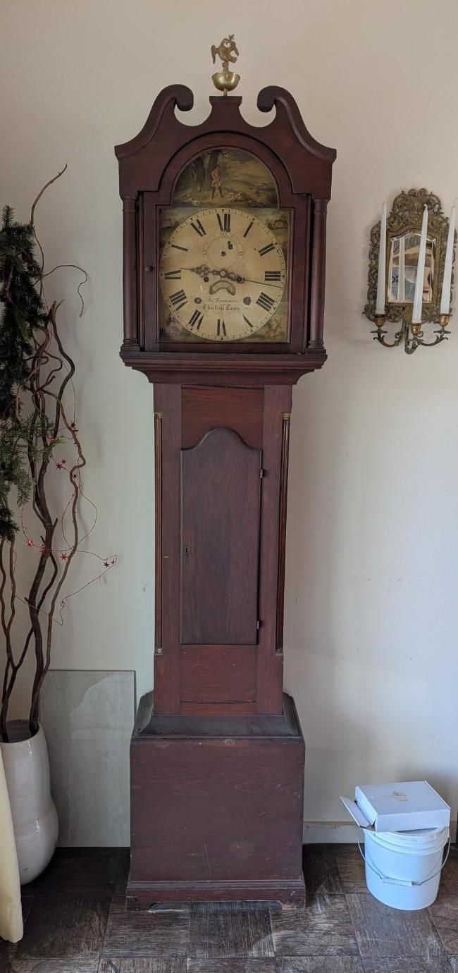 A tall, dark wood grandfather clock with a decorative dial and gold finial stands in a room next to a wall sconce.