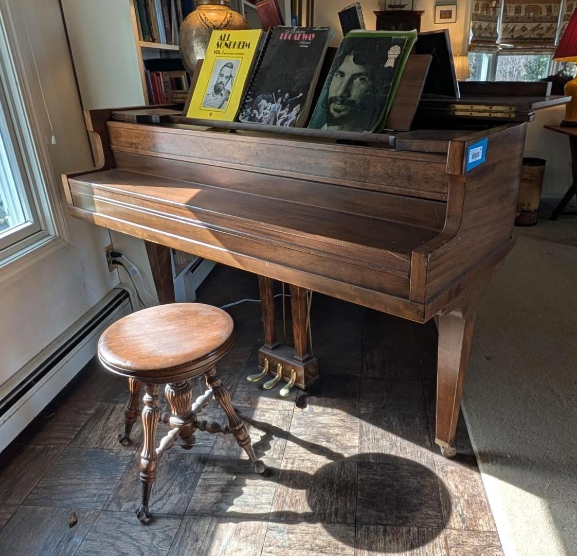 A wooden upright piano with a round stool on a tiled floor, featuring sheet music on the stand in a sunlit room.