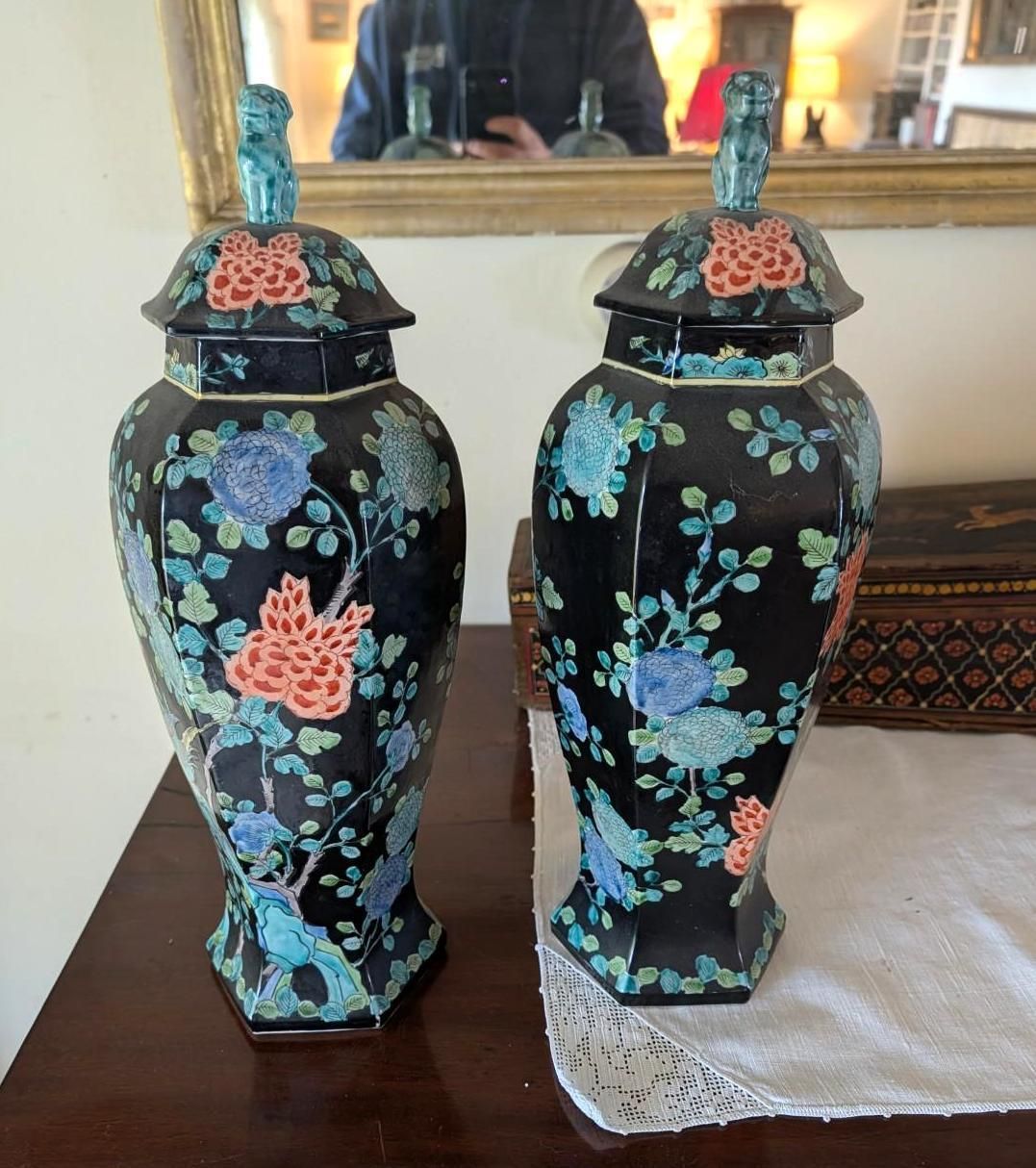 Two matching black hexagonal lidded vases decorated with vibrant floral patterns, sitting on a wooden surface.