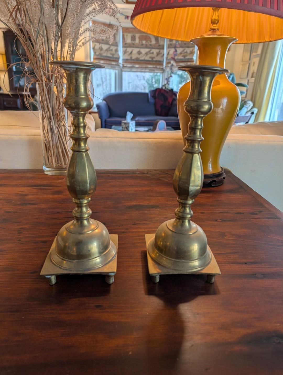 Two brass candlesticks on a wooden table, with a lamp and decorative branches visible in the background.