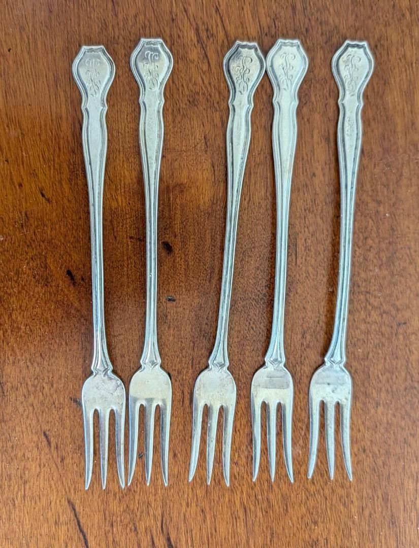 Five vintage silver cocktail forks with decorative handles arranged side-by-side on a wooden surface.