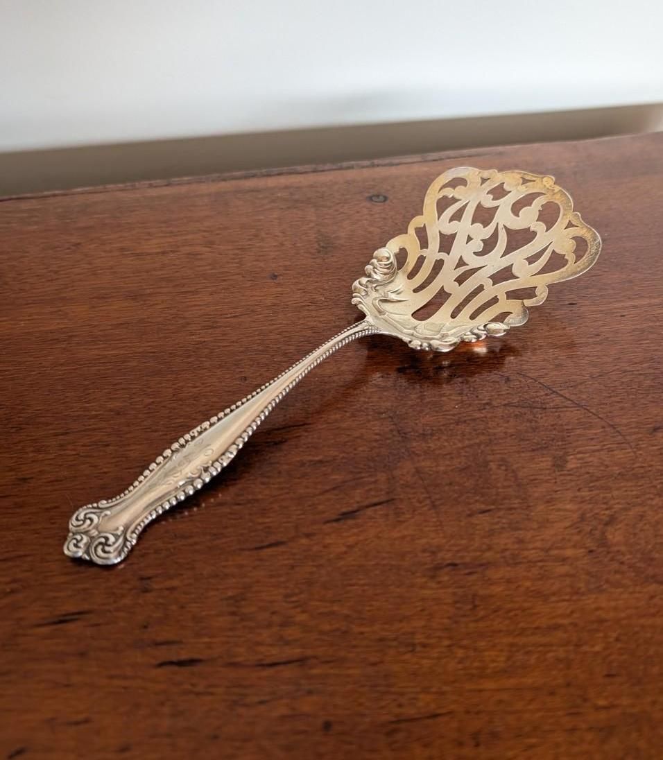 An ornate silver-plated serving spoon with a decorative, pierced, filigree bowl resting on a dark wooden surface.