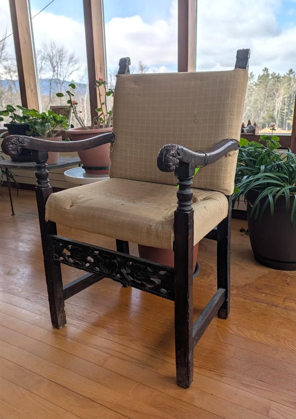 A dark wood armchair with carved armrests and a light beige, woven fabric seat and back, set in a sunlit room with plants.