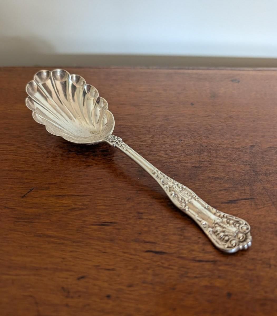 A silver, shell-shaped decorative spoon with an ornate handle, resting on a dark wooden surface.