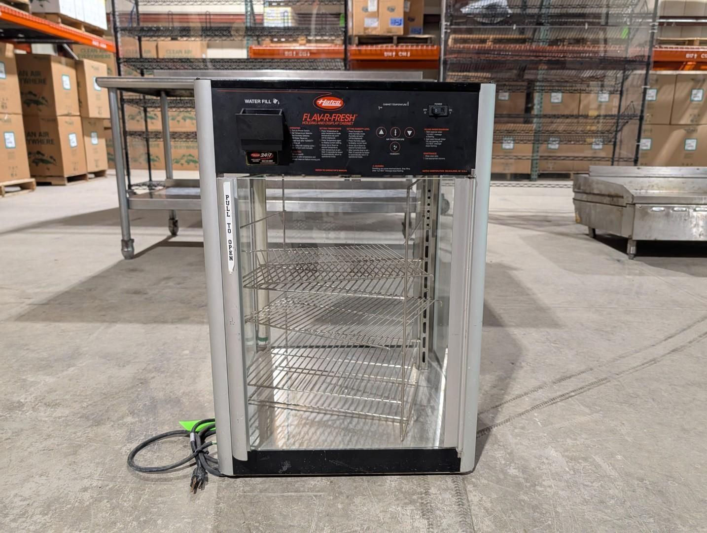 Stainless steel industrial appliance in a warehouse, with black control panel and coiled power cord