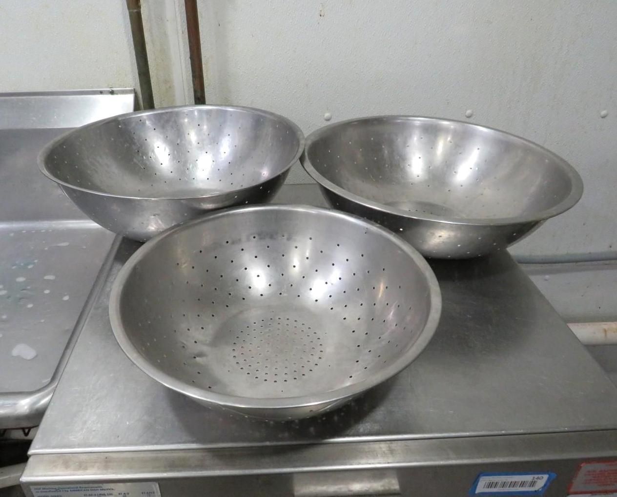 Three stainless steel colander bowls on a metal surface, likely in a kitchen setting.