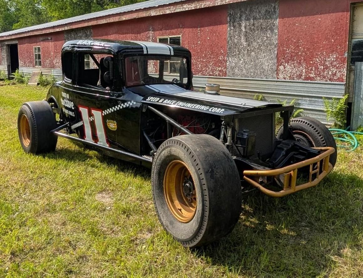 Black vintage race car with gold wheels parked on grass beside a red barn.