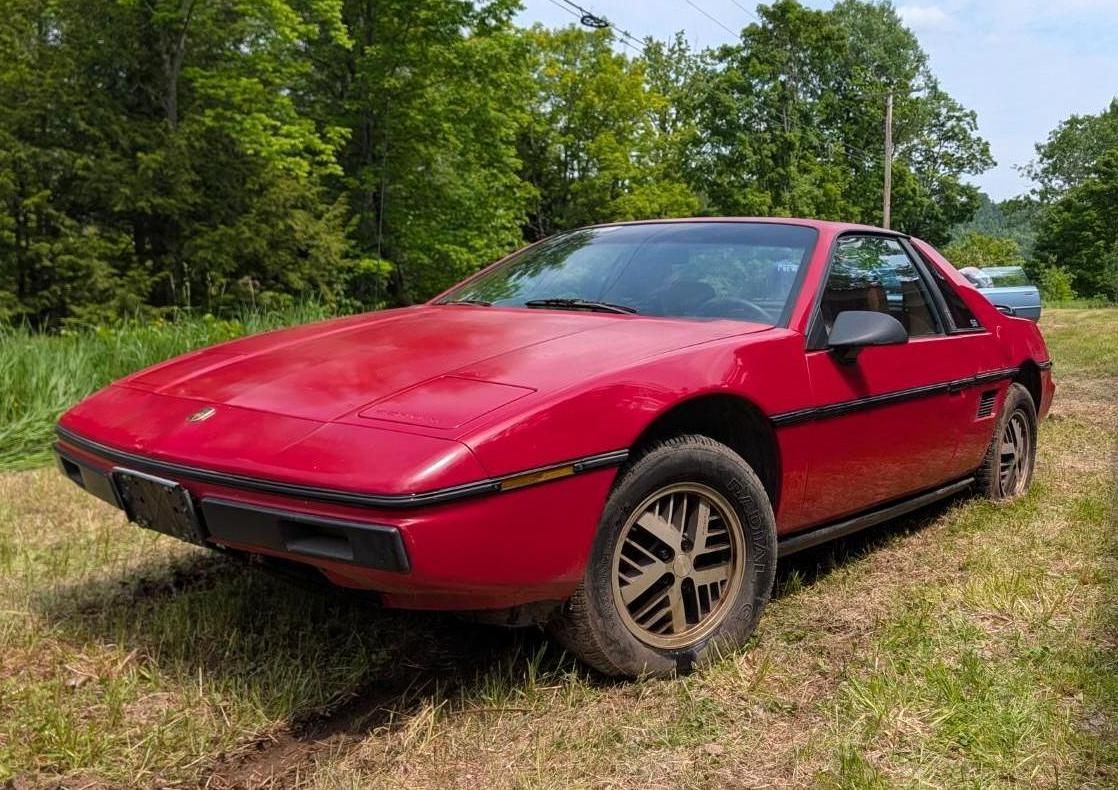 Red sports car parked on grass in a wooded area under a partly cloudy sky