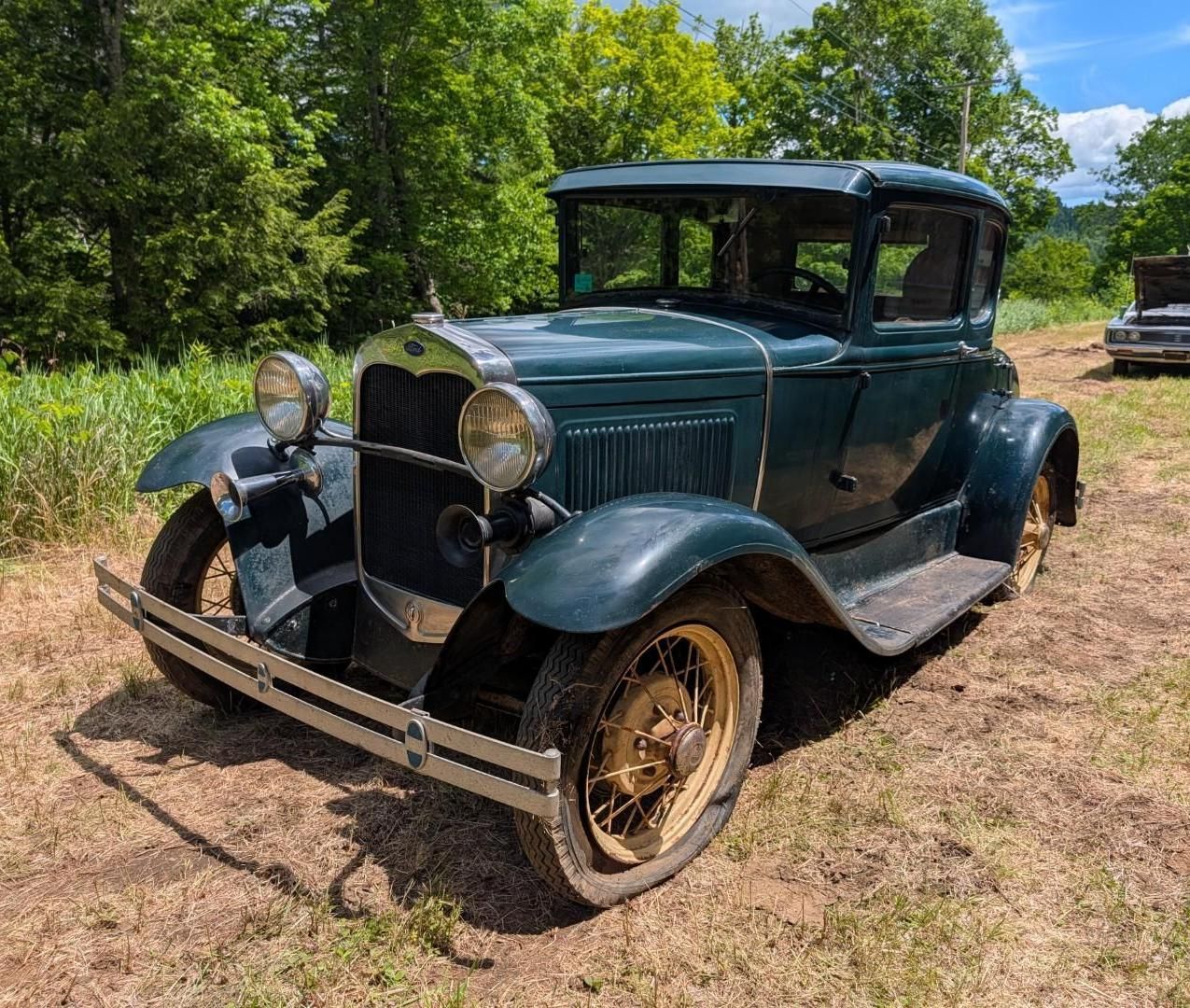 Vintage dark green car parked on dirt beside trees, with brass headlights and gold wheels