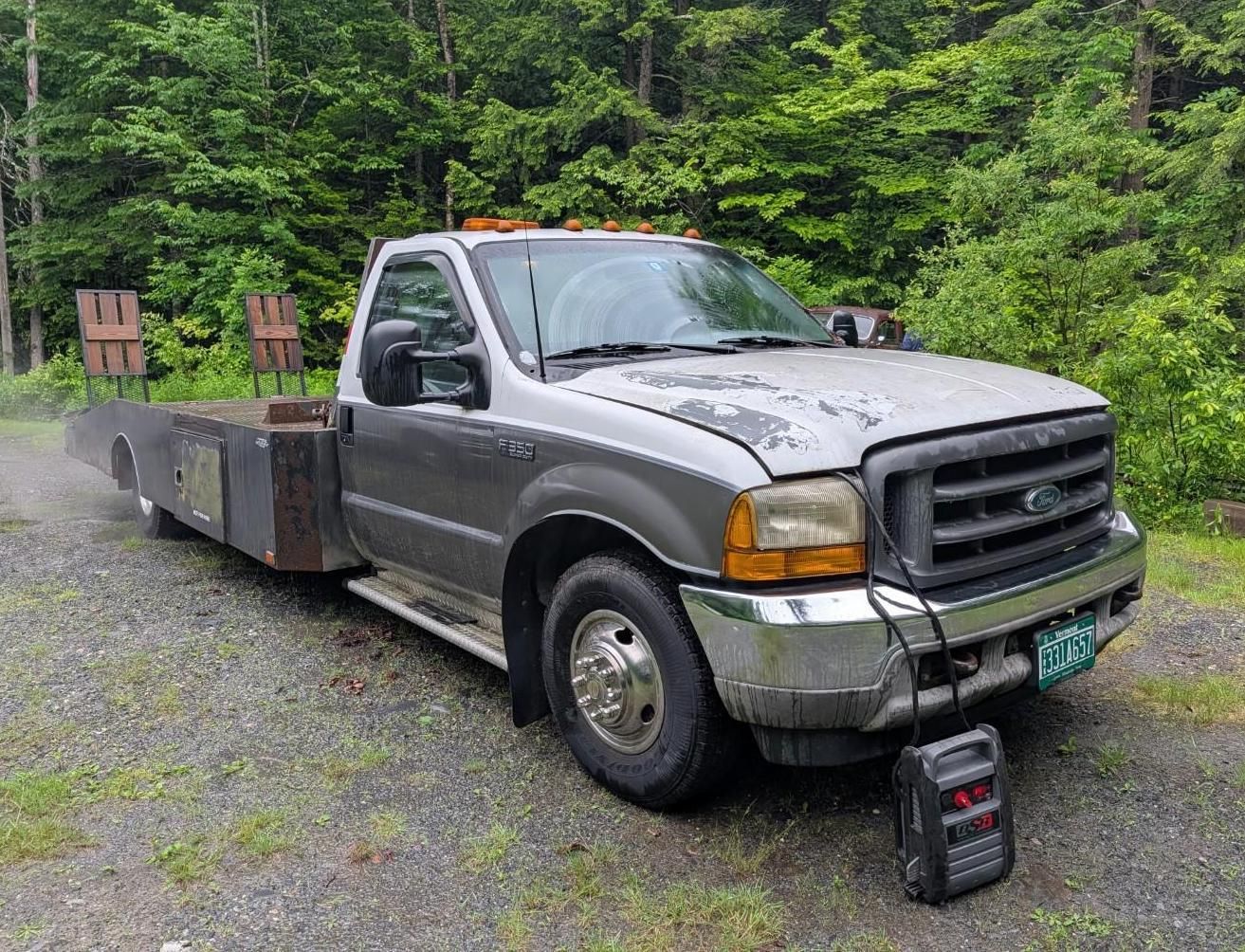 Gray flatbed tow truck parked on gravel beside a forest, with a portable battery charger in front.