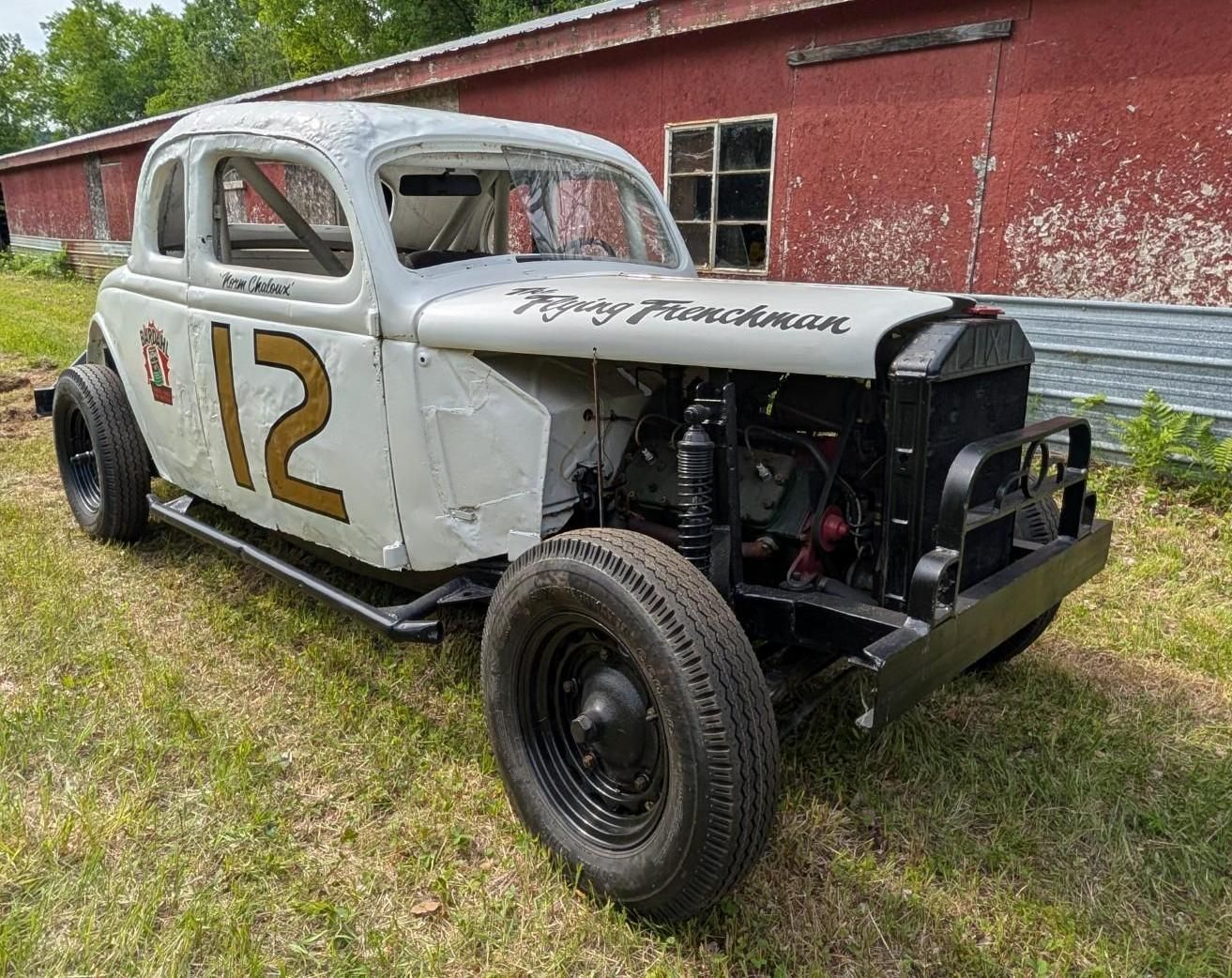 Vintage white race car with number 12, missing front bodywork, parked on grass by a red barn