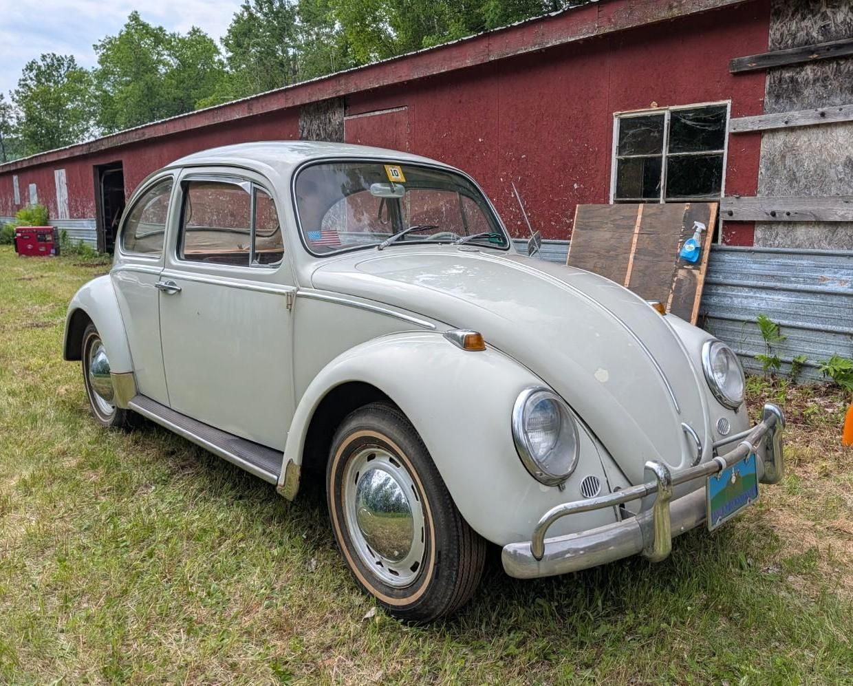 Vintage light-gray Volkswagen Beetle parked beside a red barn in a grassy yard