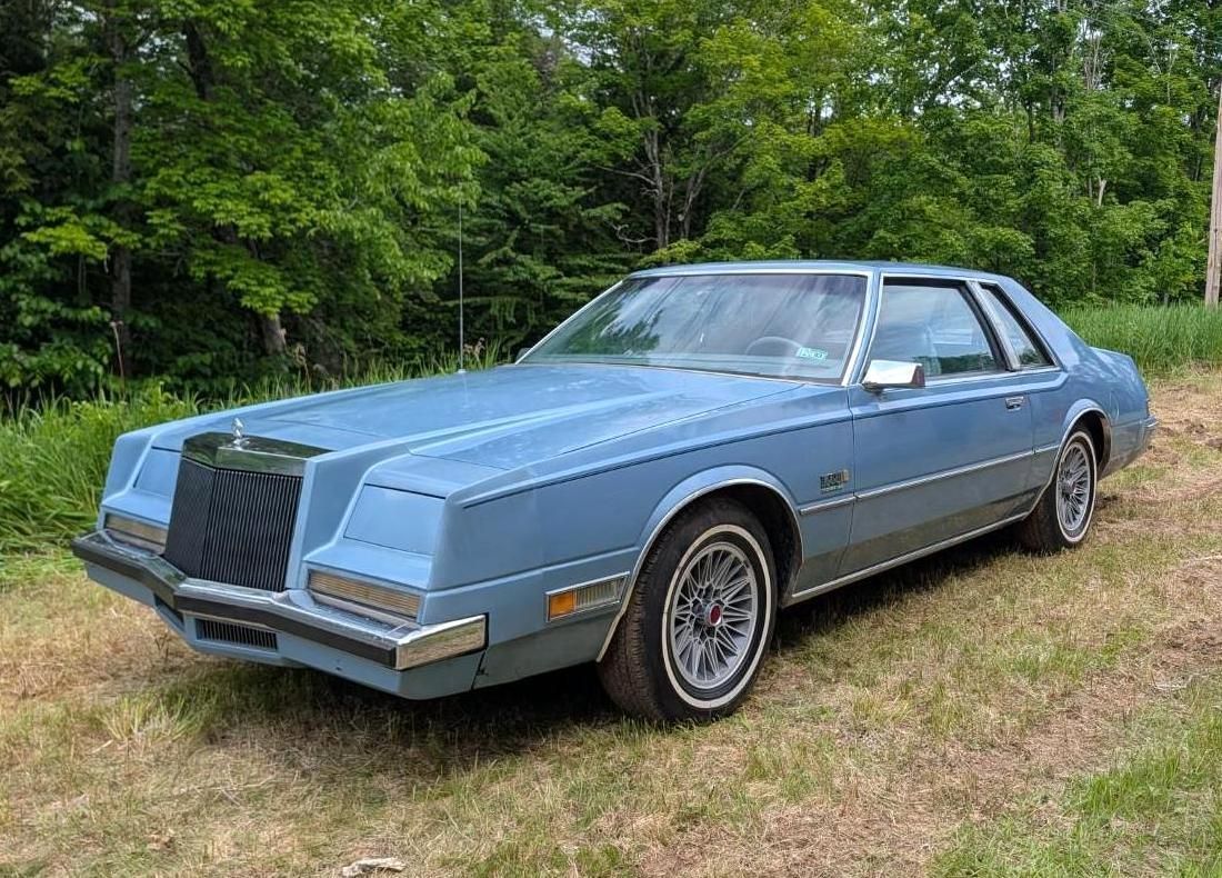 Blue vintage coupe parked on grass beside a tree line