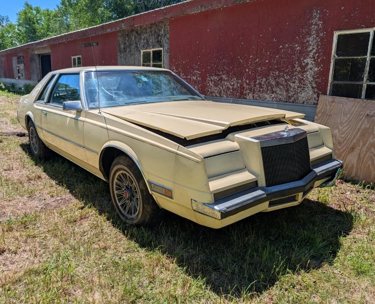 Old beige classic car parked beside a red building in a grassy lot, front hood slightly open
